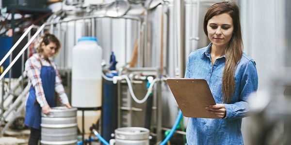A brewery worker inspects a stainless-steel tank while holding a tablet
