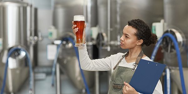 Person inspecting a glass of beer in a production facility with brewing equipment in the background and holding a clipboard